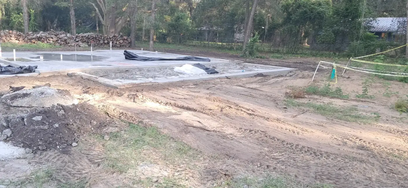 Tractor-graded soil and access around a new concrete home foundation beneath mature trees