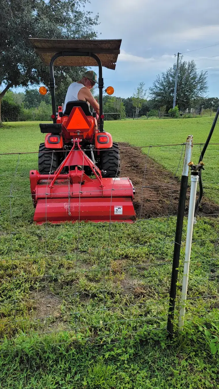 Compact tractor rototilling a straight planting strip through a grassy field behind a wire fence