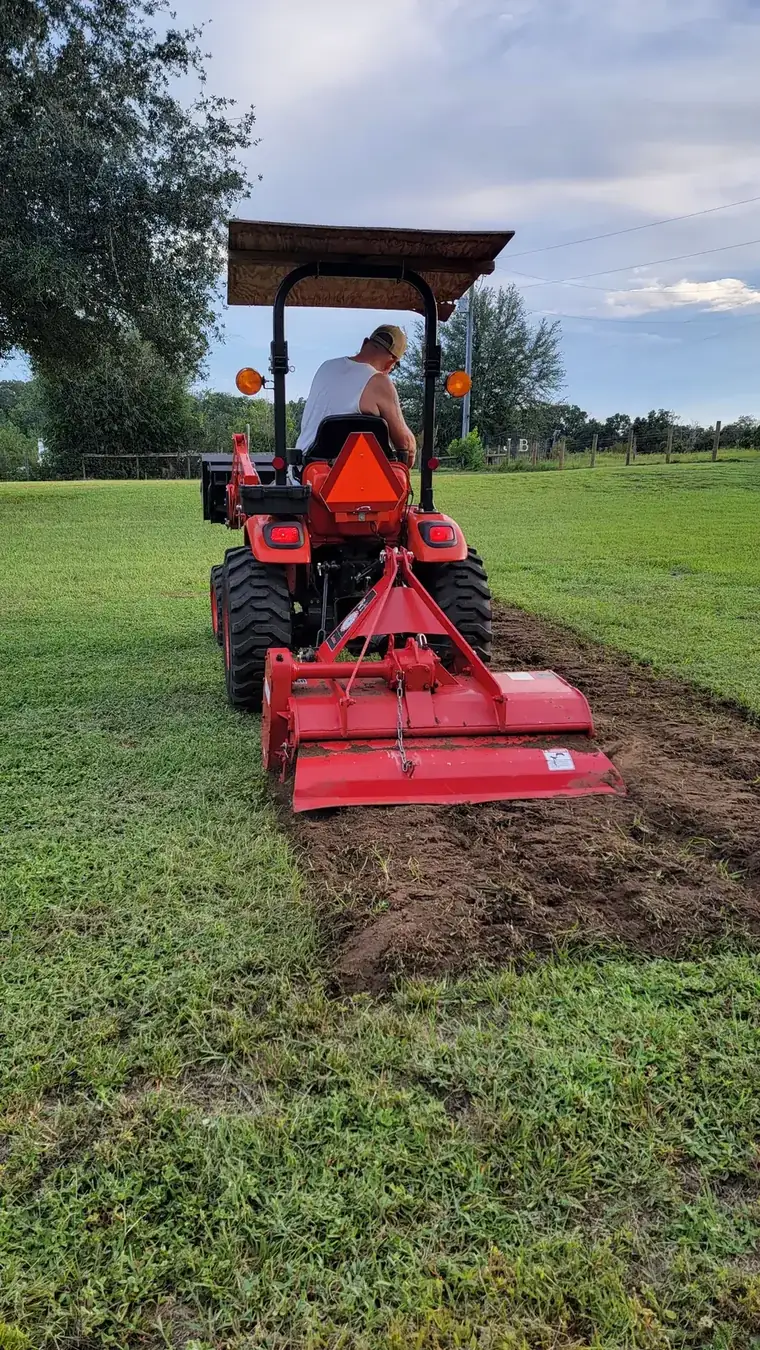 Rear tiller turning sod into loose soil as the tractor works a garden strip across the pasture