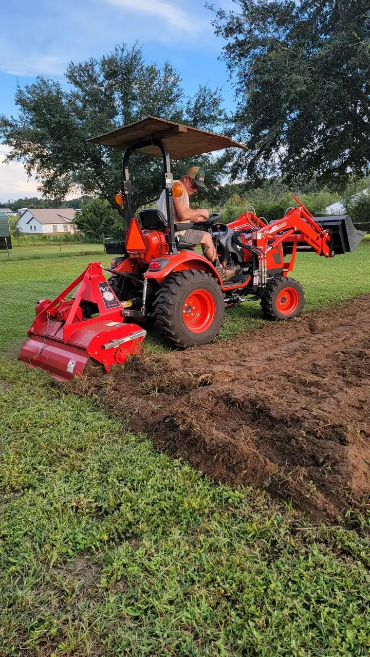 Side view of the compact tractor and rear tiller working the edge of a newly tilled strip